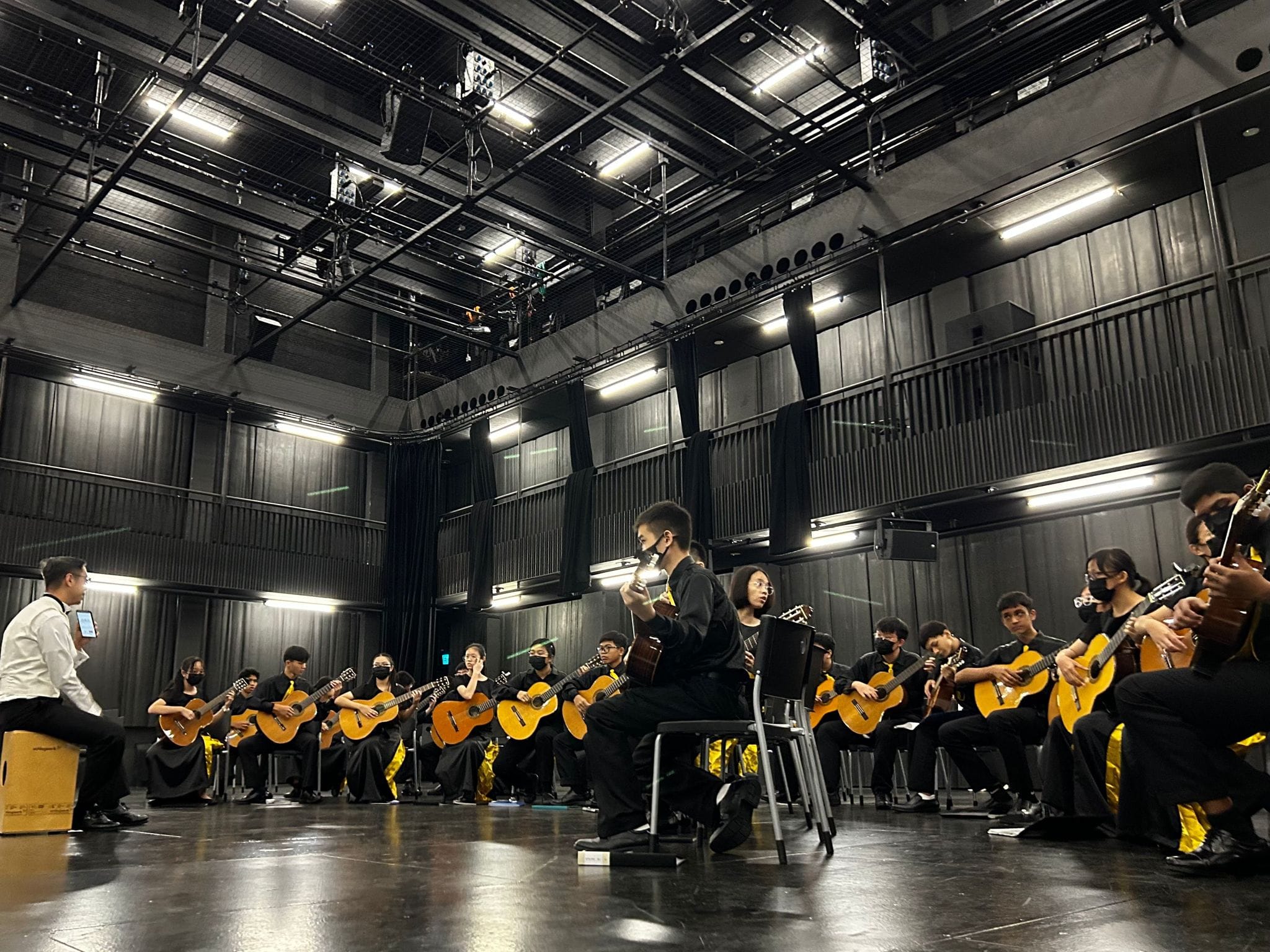 A large group of young musicians in black attire playing classical guitars on a dark indoor stage.